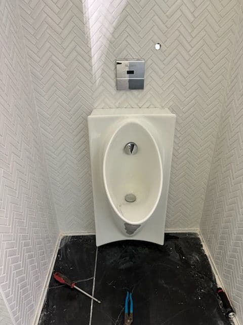 Modern wall-mounted urinal in herringbone tiled bathroom with tools nearby.