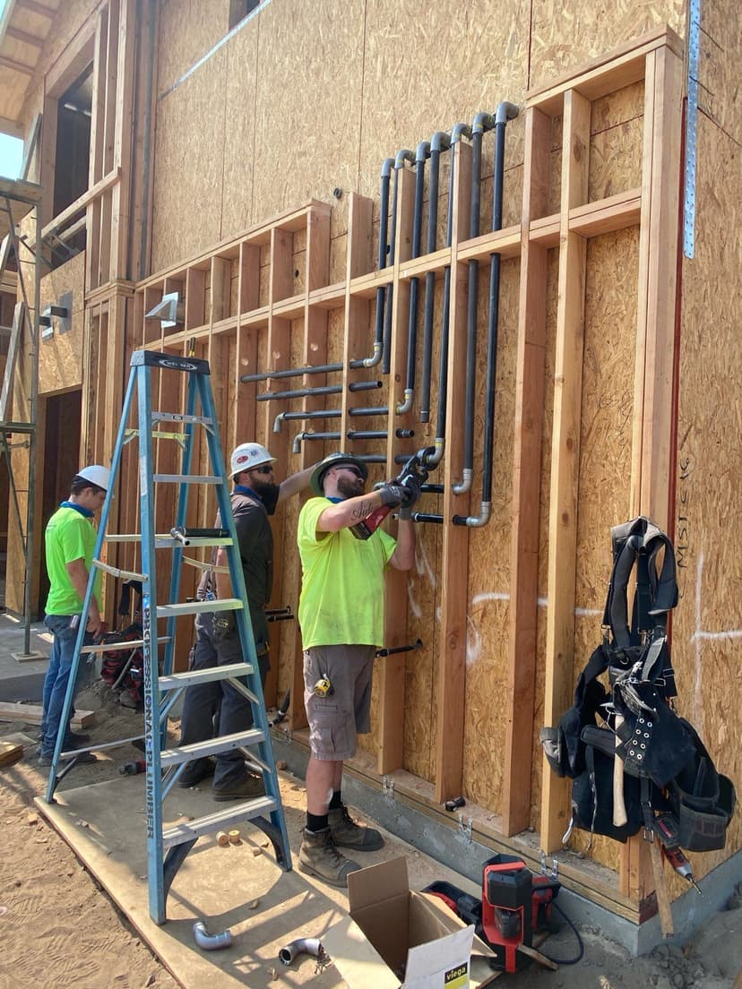 Construction workers installing plumbing on a wooden wall framework at a building site.