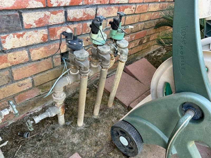 Household irrigation valves and pipes next to brick wall with garden equipment nearby.