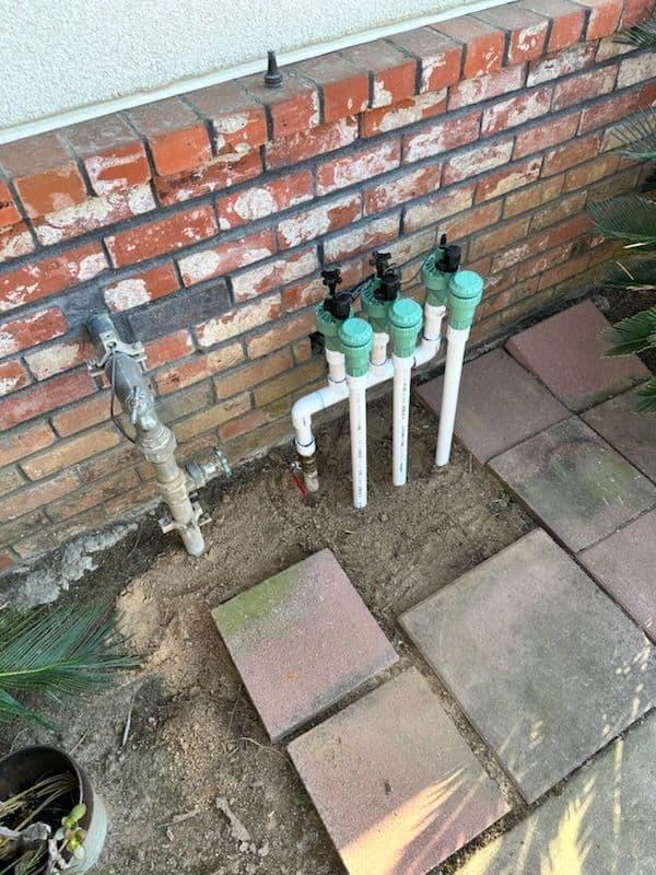 Irrigation system with PVC pipes and valves installed near a brick wall and paver stones.
