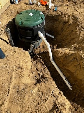 Ground installation of a green sump pump with white PVC piping in a sandy trench.