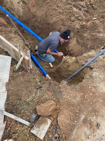 Worker digging trench for plumbing installation with blue pipes in construction site.