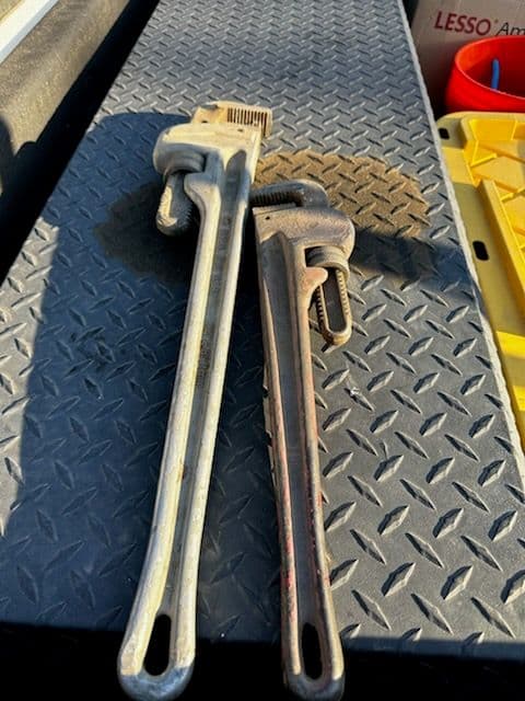 Two pipe wrenches resting on a textured metal surface, one larger than the other.