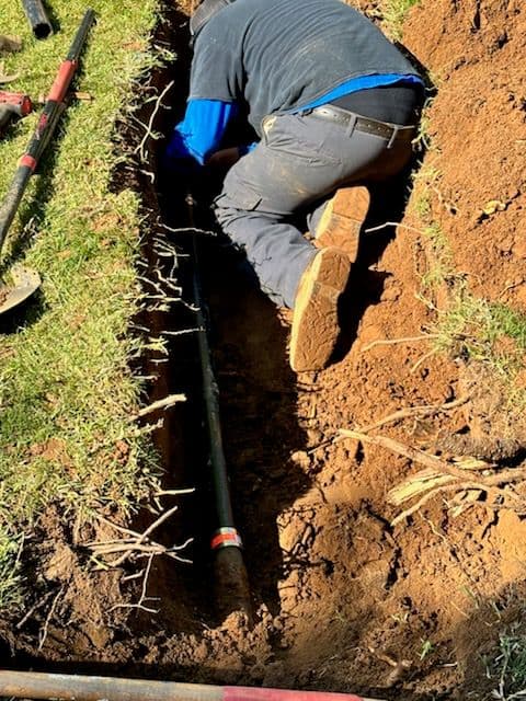 Worker installing a pipe in a trench, surrounded by soil and tools on a grassy area.