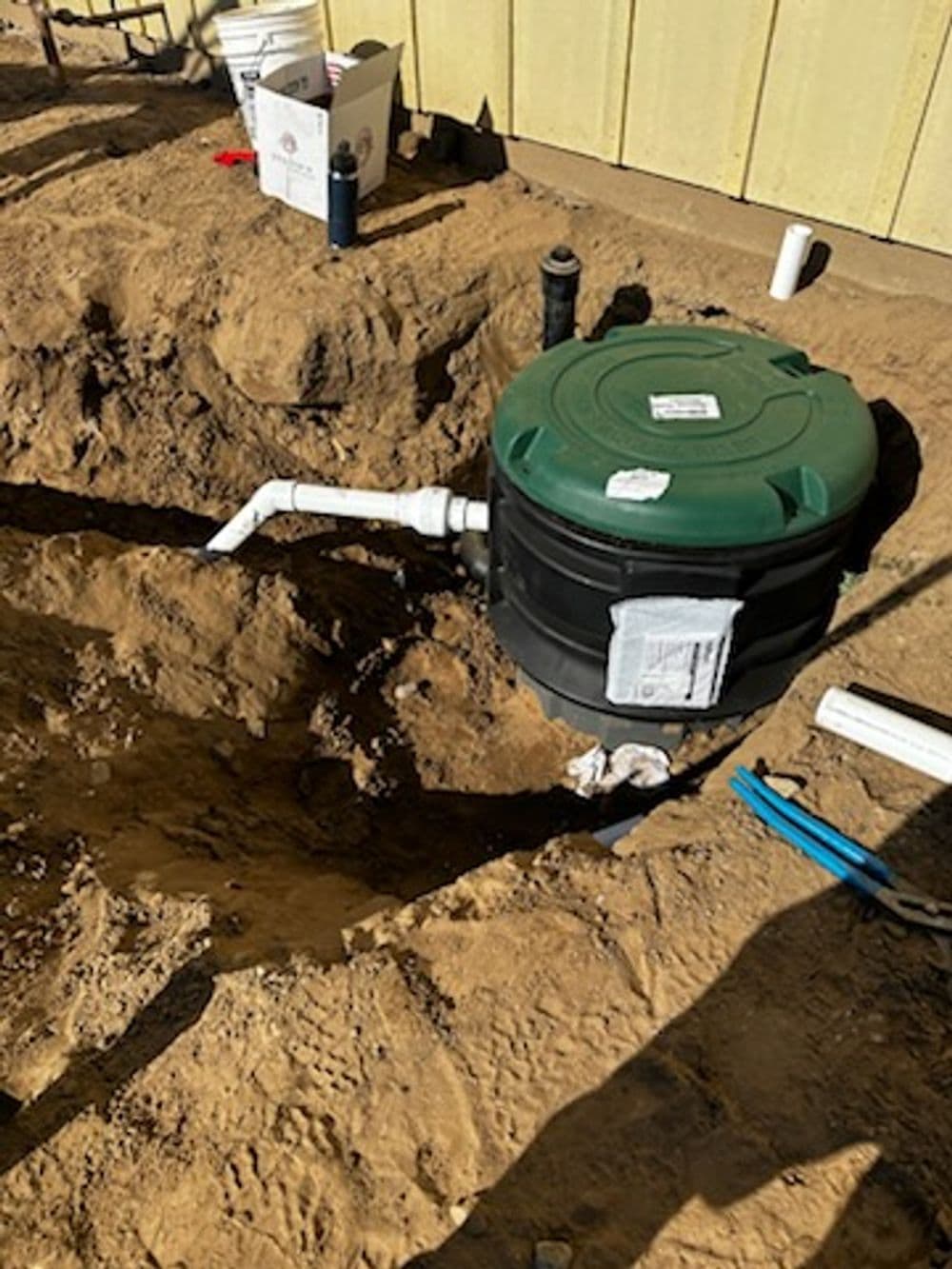 Outdoor installation of a black and green water storage tank with pipes, on sandy ground.