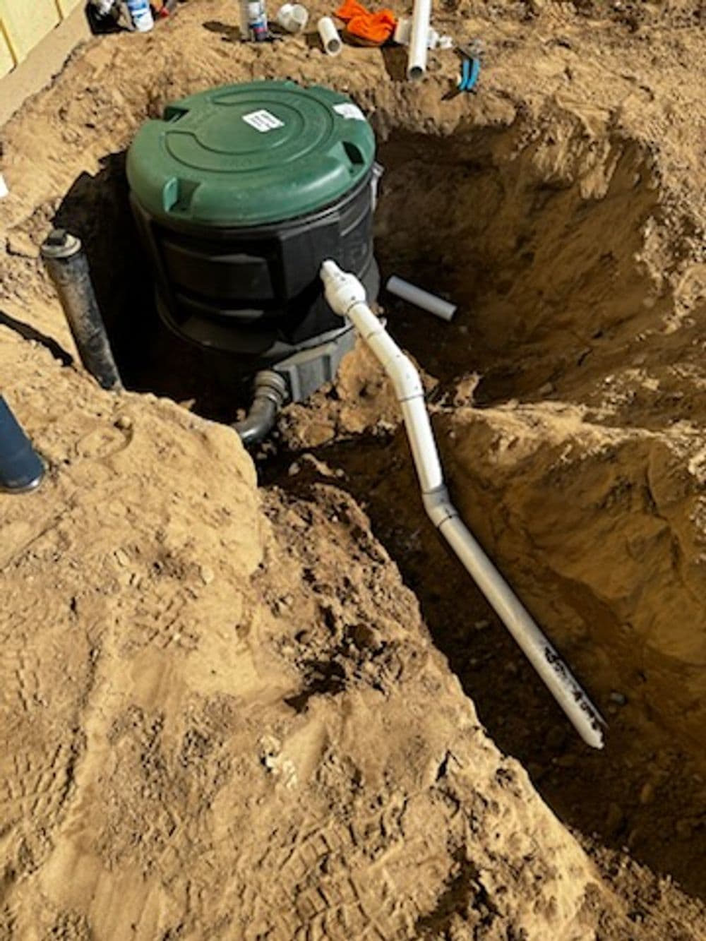 Green septic tank installed in a dug-out trench with white piping and surrounding dirt.